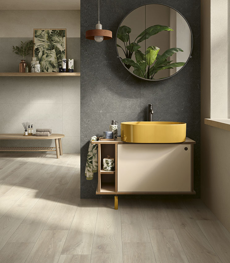 Salle de bains contemporaine avec des surfaces des collections Surround Outline Soft, Draft et Borealis Inari, caractérisée par un mur sombre, une vasque ovale jaune, un meuble suspendu, un grand miroir rond, des plantes décoratives et un sol clair effet bois.