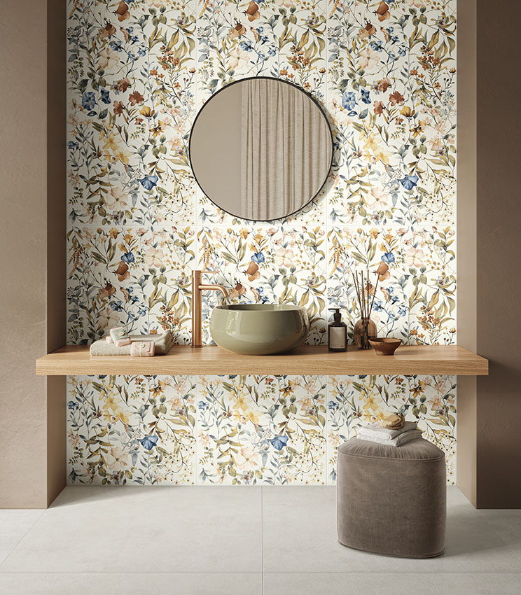 Salle de bains contemporaine avec des surfaces des collections La Matière Talc et La Fleur, caractérisée par un revêtement floral aux tons naturels, une vasque vert olive, une tablette en bois, un miroir rond et un pouf rembourré sur un sol clair.