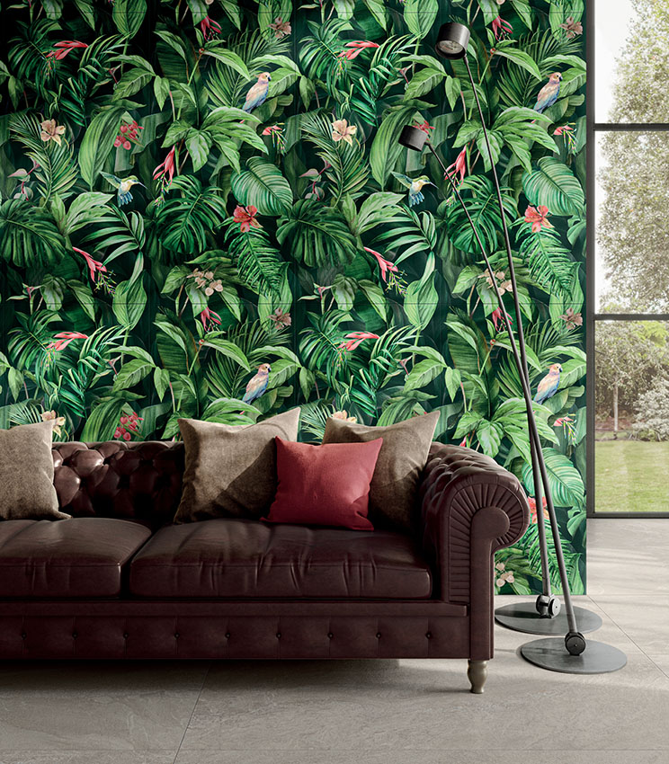 Salon contemporain avec un revêtement mural décoratif de la collection Glam Vision, caractérisé par un motif tropical de feuilles, de fleurs et d’oiseaux, un canapé en cuir foncé avec des coussins aux tons neutres et bordeaux, des lampadaires noirs et une grande baie vitrée donnant sur le jardin.