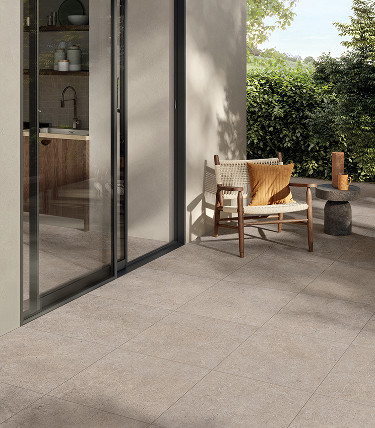 Terrasse contemporaine avec un sol de la collection Surround Row, caractérisé par de grandes dalles effet pierre dans des tons beige, un fauteuil en bois et corde, une table d’appoint sculpturale et une végétation luxuriante à l’arrière-plan. De grandes baies vitrées relient l’espace extérieur à la cuisine intérieure.