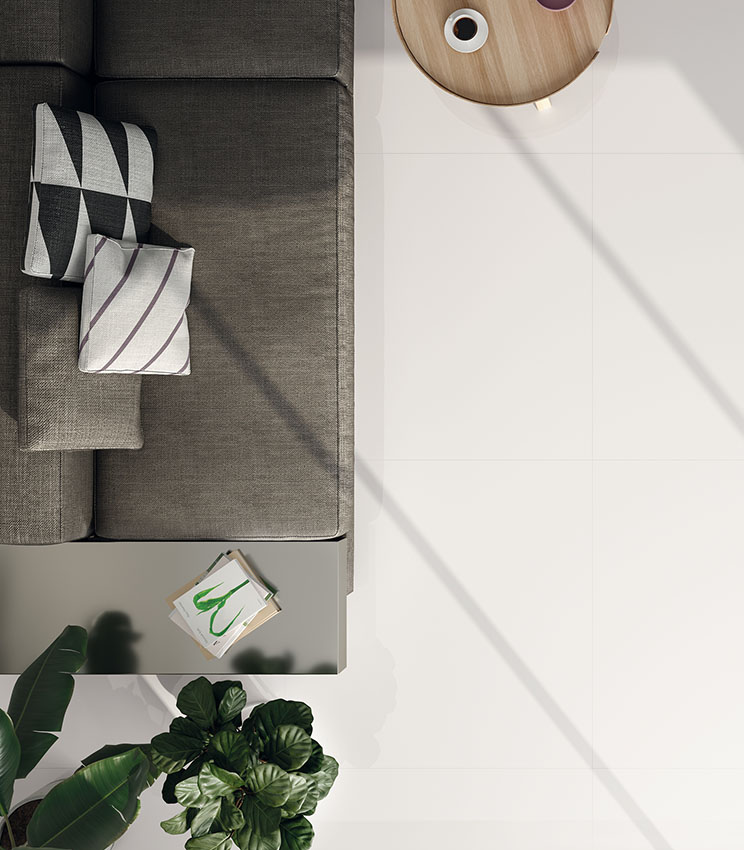 Vue de dessus d’un salon moderne avec canapé gris, table basse ronde en bois clair et sol brillant effet marbre blanc.