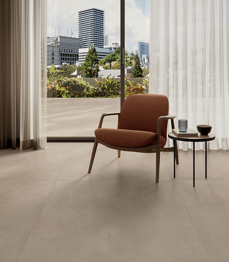 Vue sur un salon moderne avec fauteuil et porte coulissante ouvrant sur une terrasse avec vue sur la ville. Sol effet béton de Panaria Ceramica, collection La Matière.