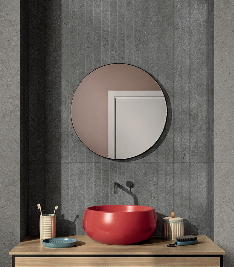 Salle de bains revêtue de carreaux en grès cérame effet pierre, avec douche à l’italienne, miroir rond et vasque à poser rouge.