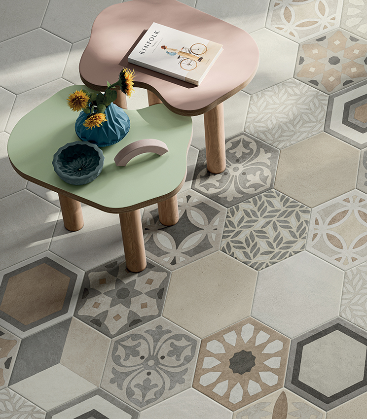 Détail d’un sol avec des surfaces des collections La Matière Hexa Talc Sable et Hexa Chic, composé de carreaux hexagonaux aux tons neutres associés à des motifs décoratifs géométriques et floraux. Deux petites tables aux formes douces, verte et rose, complètent la scène avec des vases et de petits objets décoratifs.
