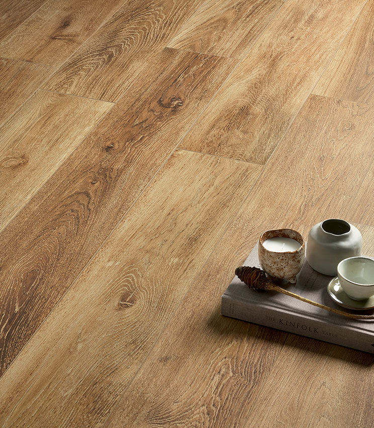 Détail d’un sol de la collection Kairos Ilios, caractérisé par des surfaces effet bois aux veinures marquées et aux tons chauds et naturels. La scène est complétée par un livre, une bougie, un petit vase et une tasse en céramique.