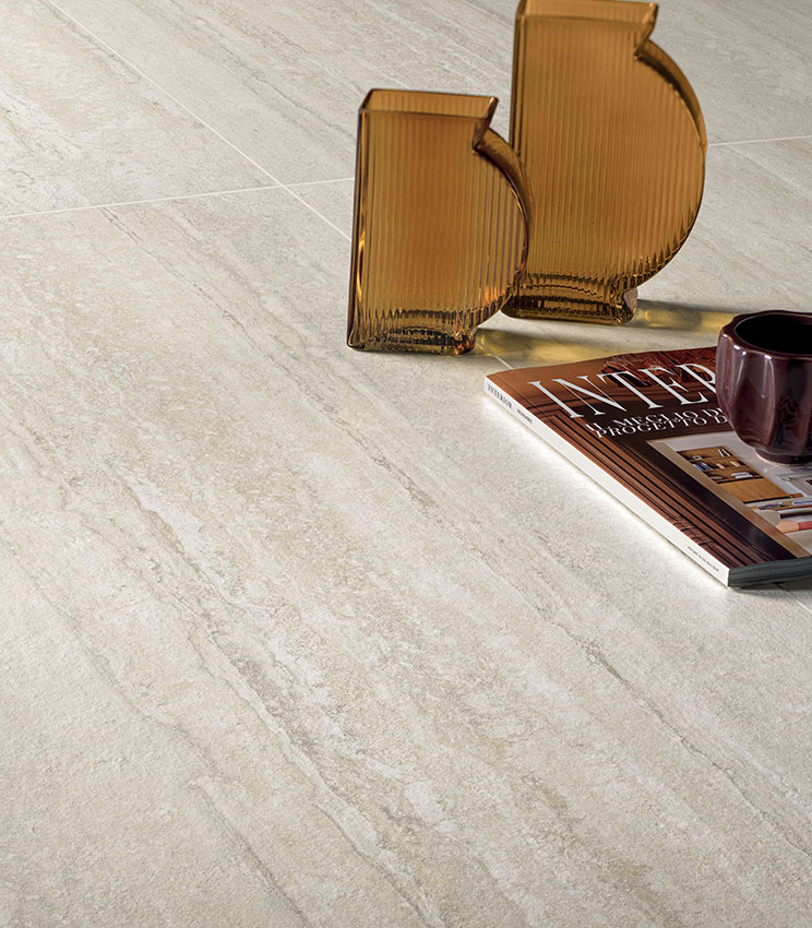 Détail d’un sol de la collection Perpetual Travertino Ivory, caractérisé par des surfaces effet travertin dans des tons clairs de beige et d’ivoire. La scène est complétée par deux vases en verre ambré, un magazine et une tasse foncée.