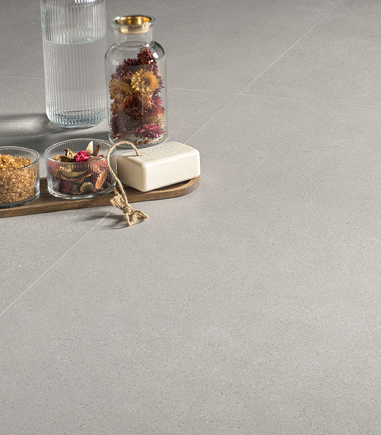 Détail d’un sol de la collection Formula Form Ash, caractérisé par une surface claire et uniforme à l’aspect texturé. La scène est complétée par une composition avec une bouteille en verre, un vase de fleurs séchées, de petits contenants et un savon posé sur un plateau en bois.