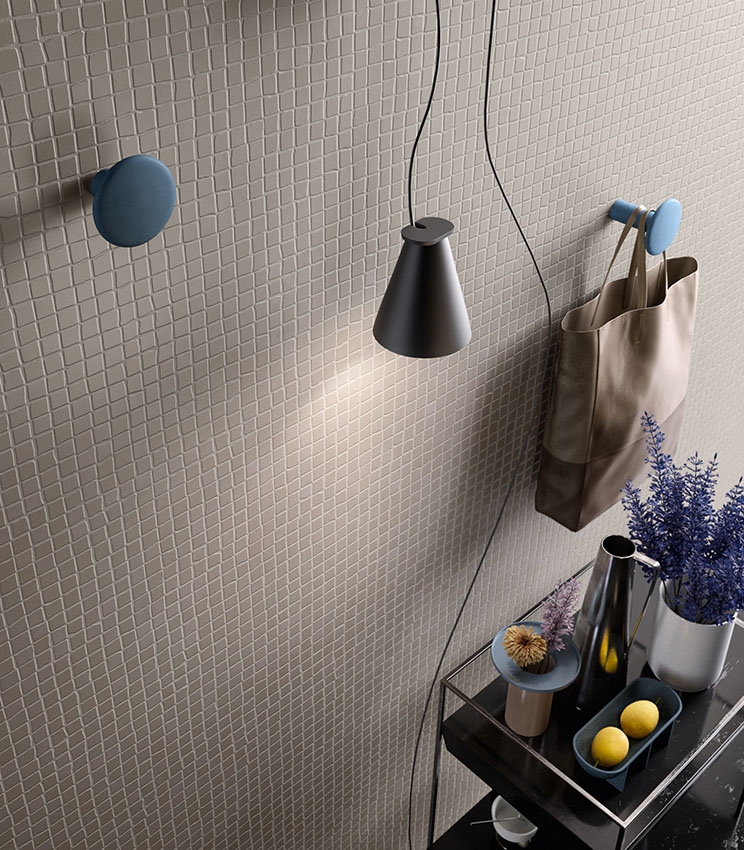 Détail d’un revêtement mural de la collection Even Dove Mosaico, caractérisé par un motif régulier de petits carreaux carrés dans des tons clairs. La scène est complétée par des patères bleues, une suspension noire, un sac en tissu et une console avec des vases, des fleurs et de petits objets décoratifs.