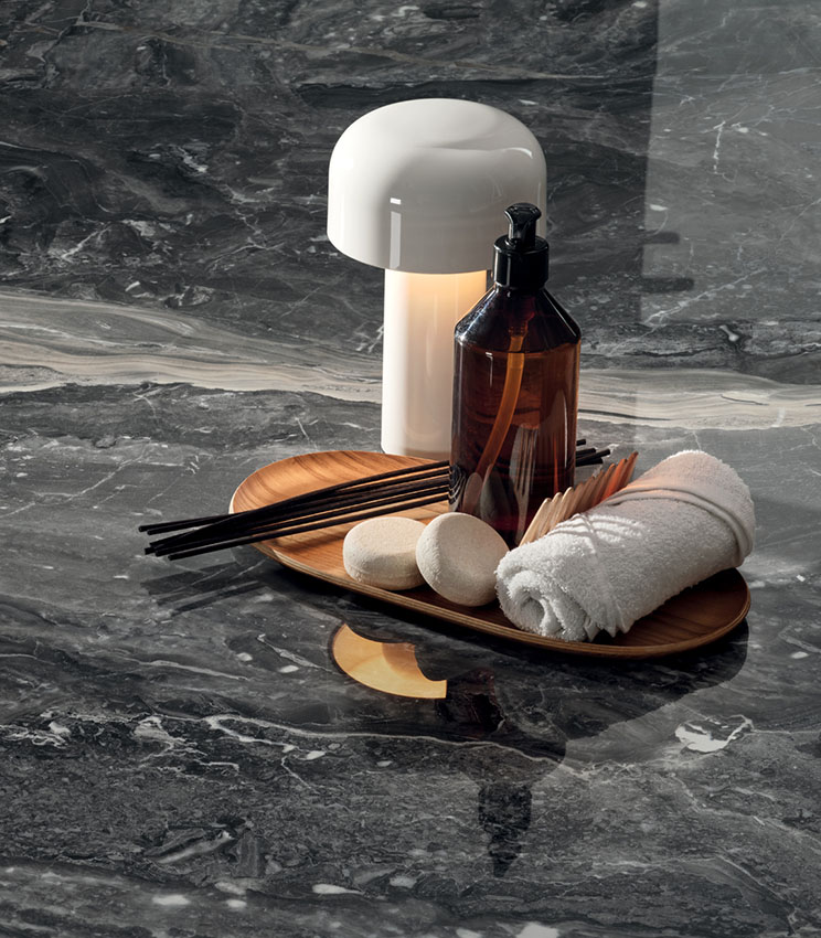 Détail d’une surface de la collection Perpetual Orobico Blend Lux, caractérisée par un effet marbre foncé aux veines claires et à la finition brillante. La scène est complétée par un plateau en bois avec un flacon, une serviette roulée, de petits accessoires et une lampe de table blanche.