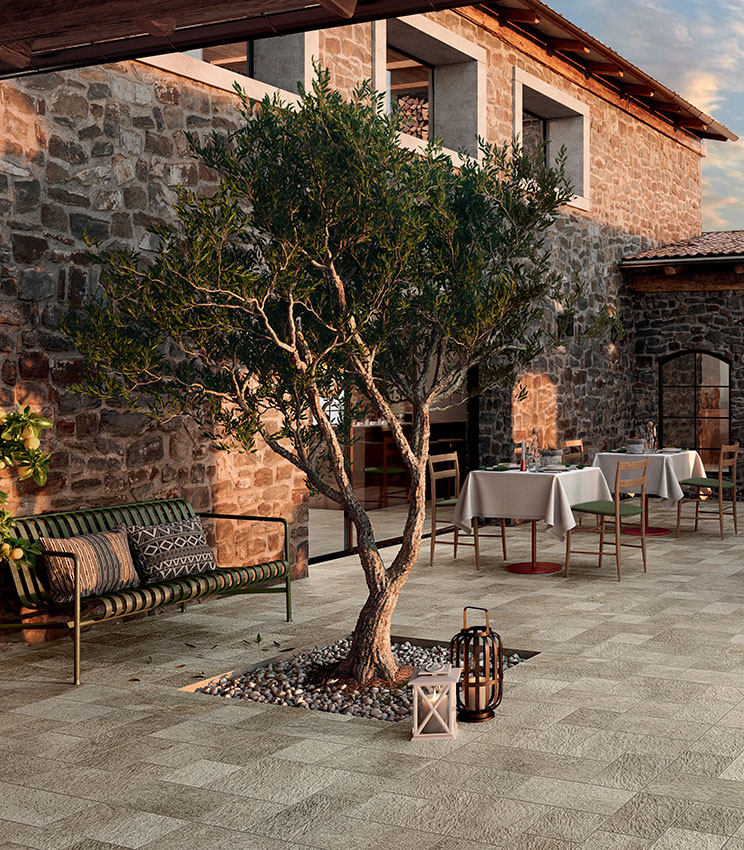 Terrasse de restaurant avec un sol de la collection Pierre des Rêves Aurore, caractérisé par des surfaces effet pierre aux tons naturels. Entouré de murs en pierre, l’espace est aménagé avec des tables et des chaises en bois, un banc, des lanternes décoratives et un olivier au centre.