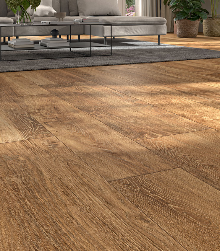 Salon contemporain avec un sol de la collection Kairos Ilios, caractérisé par des surfaces effet bois aux tons chauds et aux veinures marquées. L’espace est aménagé avec un grand canapé gris, une table basse en métal noir, un tapis foncé et des plantes décoratives éclairées par la lumière naturelle.