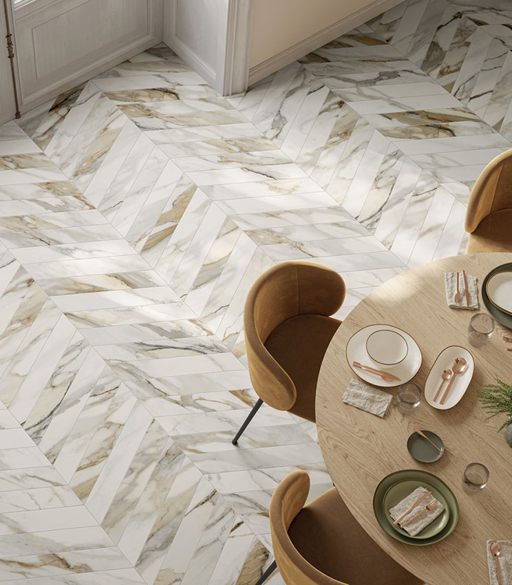 Salle à manger contemporaine avec un sol de la collection Perpetual Venato Gold Soft Chevron, caractérisé par un motif chevron effet marbre blanc aux veinures grises et dorées. L’espace est aménagé avec une table ronde en bois clair, des chaises rembourrées couleur ocre et une mise en table dans des tons neutres.