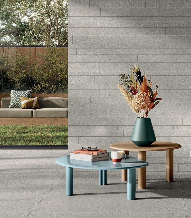 Salon contemporain avec des surfaces des collections Surround Shape et Shape Muretto Soft, caractérisé par un sol effet pierre grise et un mur revêtu d’éléments rectangulaires coordonnés. L’espace est aménagé avec des tables basses aux lignes épurées, un vase avec une composition florale et une large ouverture sur un jardin avec canapé extérieur.