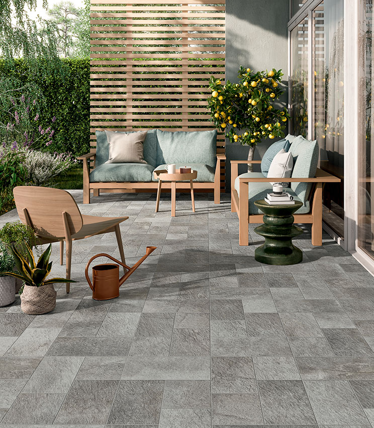 Terrasse contemporaine avec un sol de la collection Pierre des Rêves Nuage, caractérisé par des surfaces effet pierre grise en formats modulaires. L’espace est aménagé avec des canapés et fauteuils en bois, des coussins vert sauge, des tables d’appoint décoratives, des plantes en pot et un agrume, au cœur d’un jardin luxuriant.