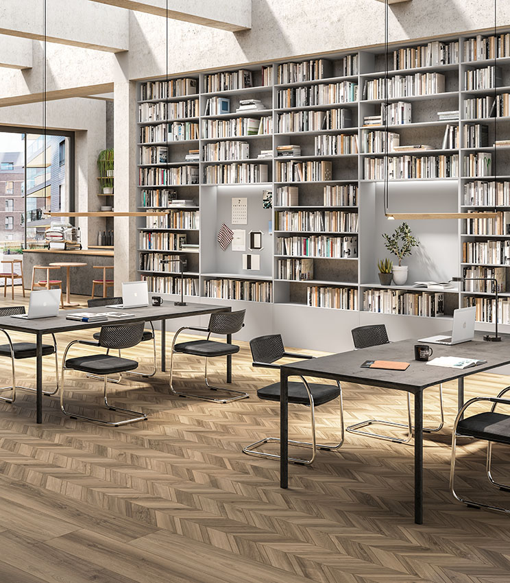 Bibliothèque et espace de travail contemporains avec un sol des collections Nuance Cendre et Nuance Cendre Chevron, caractérisé par des surfaces effet bois aux tons naturels et une pose géométrique. L’espace comprend de grandes bibliothèques murales, des tables de travail avec des chaises noires, des ordinateurs portables, des plantes décoratives et de grandes fenêtres apportant beaucoup de lumière naturelle.