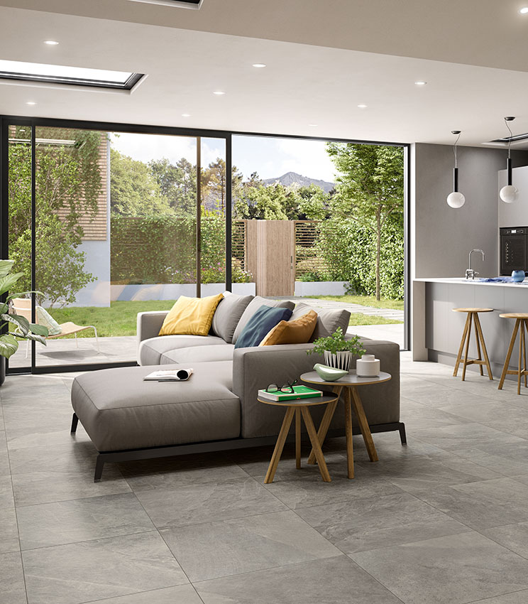 Vue d'un espace ouvert salon-cuisine avec une grande porte vitrée donnant sur un petit jardin privé. Sol en grès cérame effet pierre grise