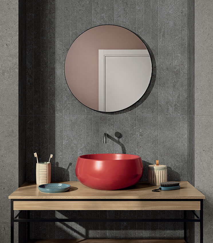 Salle de bains contemporaine avec des surfaces des collections Surround Shape Soft et Outline Carving, caractérisée par des revêtements gris effet pierre et une zone centrale à motif linéaire vertical. L’espace comprend une vasque rouge brillante sur un plan en bois, un miroir rond, une robinetterie foncée et de petits accessoires de salle de bains.