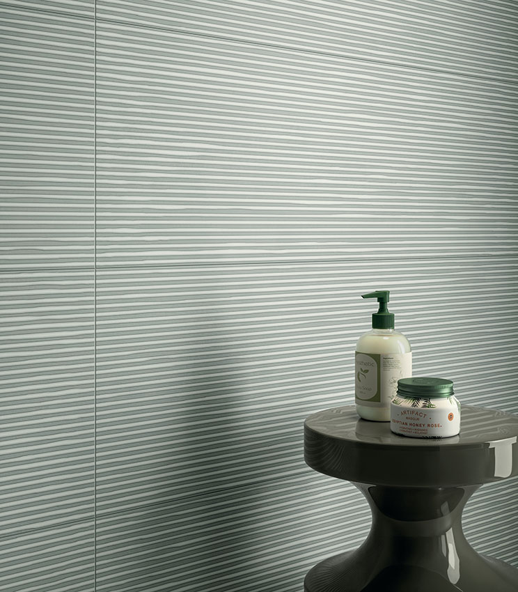 Détail d’une salle de bains contemporaine avec un revêtement mural de la collection Lullaby Linear Green, caractérisé par une surface vert clair à motif linéaire horizontal. La scène est complétée par une petite table sombre aux formes sculpturales avec des flacons et de petits produits de salle de bains.