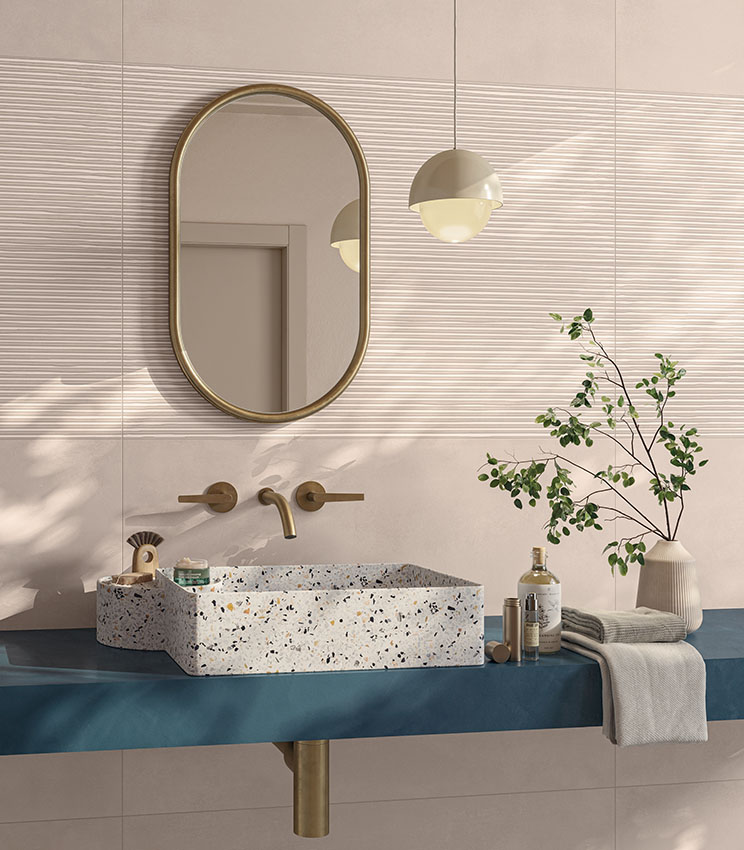 Salle de bains contemporaine avec des surfaces des collections Lullaby Fairytale Blush et Linear Blush, caractérisée par des revêtements rose poudré avec des zones à relief linéaire. L’espace comprend une vasque rectangulaire effet terrazzo, un plan bleu, un miroir ovale à cadre doré, une robinetterie assortie, une suspension et des branches vertes décoratives.