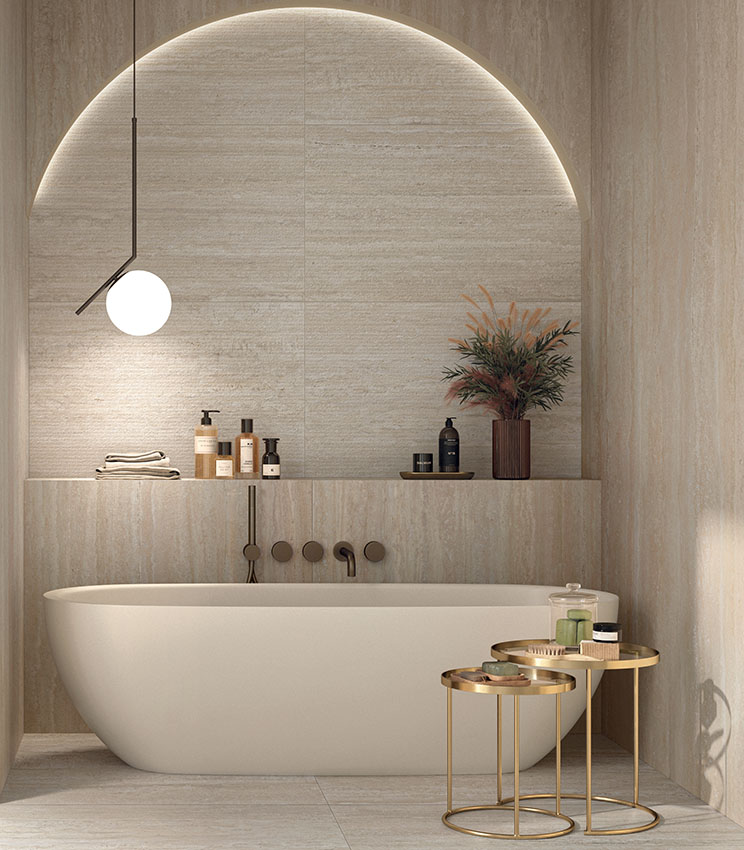 Salle de bains contemporaine avec des surfaces des collections Perpetual Travertino Ivory Edge, Travertino Ivory et Travertino Cream, caractérisées par un effet travertin dans des tons chauds et naturels. L’espace comprend une baignoire îlot blanche intégrée dans une niche en arche rétroéclairée, des étagères intégrées, une suspension et des tables d’appoint en métal doré avec des accessoires de salle de bains.