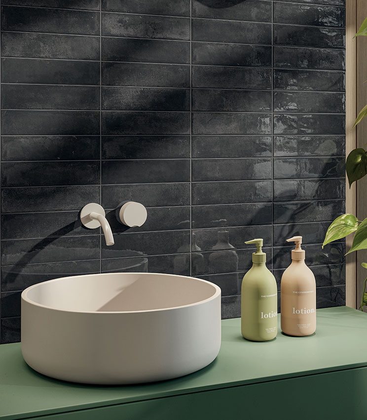 Détail d’une salle de bains contemporaine avec un revêtement mural de la collection Workshop Core, composé de carreaux rectangulaires foncés à la finition brillante et irrégulière. La scène comprend une vasque blanche posée sur un meuble vert, une robinetterie murale, des flacons coordonnés et une plante décorative.