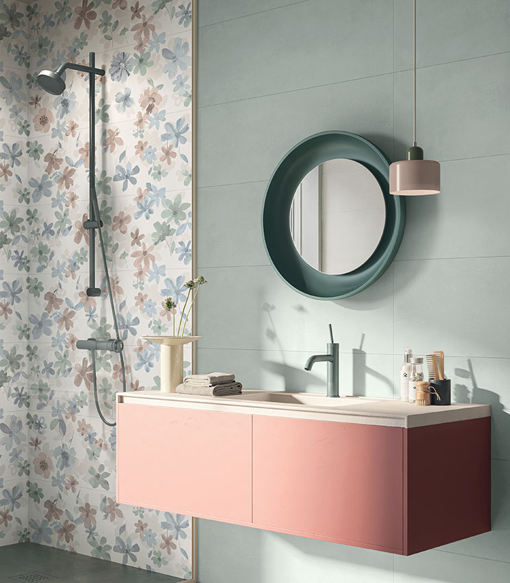 Salle de bains contemporaine avec des surfaces des collections Lullaby Silent Green, Florè et Borealis Abisko, caractérisée par des revêtements vert clair et un mur décoratif à motif floral dans des tons pastel. L’espace comprend un meuble vasque suspendu rose, un miroir rond vert foncé, une robinetterie coordonnée, une suspension et un espace douche avec colonne noire.