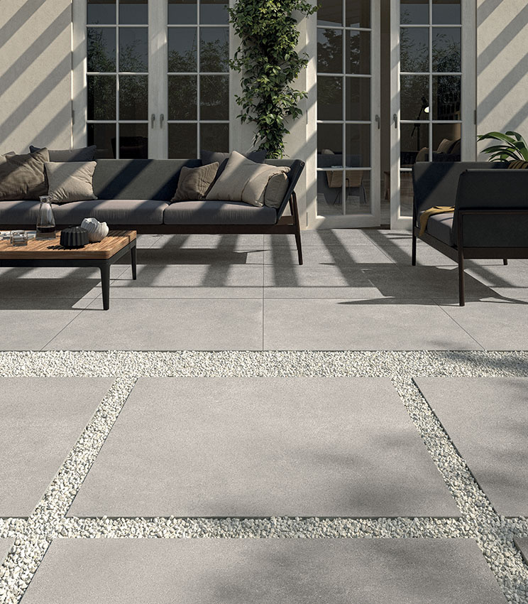 Vue d’un patio aménagé avec un salon moderne composé d’un canapé et d’un fauteuil. Sol en grès cérame de 20 mm effet béton, posé par collage. Au premier plan, un cheminement extérieur réalisé avec de grandes dalles céramiques posées à sec sur du gravier. | Collection Formula de Panaria Ceramica