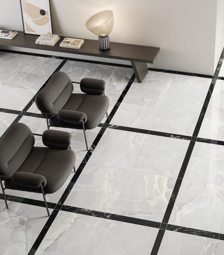 Vue en hauteur d'un vaste salon lumineux avec un sol en grès cérame effet marbre noir et blanc posé en tapis encadré, avec une paire de fauteuils sombres et un meuble-banc appuyé contre une cloison blanche.
