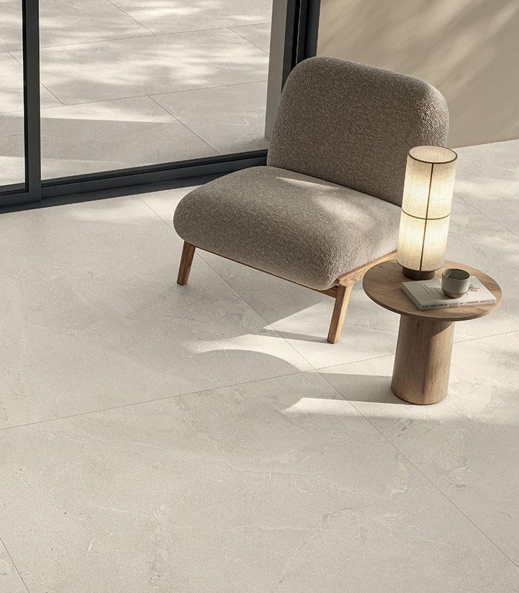 Espace de vie moderne et lumineux avec fauteuil rembourré, table d’appoint en bois et sol en grès cérame effet pierre claire, en continuité avec l’extérieur.