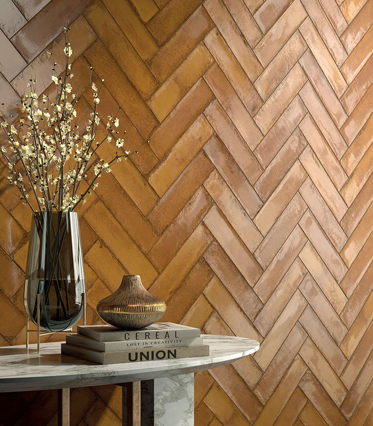 Détail d’un mur revêtu avec la collection Workshop Spice, caractérisé par des éléments rectangulaires posés en chevrons dans des tons chauds de terre cuite. Au premier plan, une table en marbre avec des livres, des vases décoratifs et des branches fleuries complète l’ambiance.