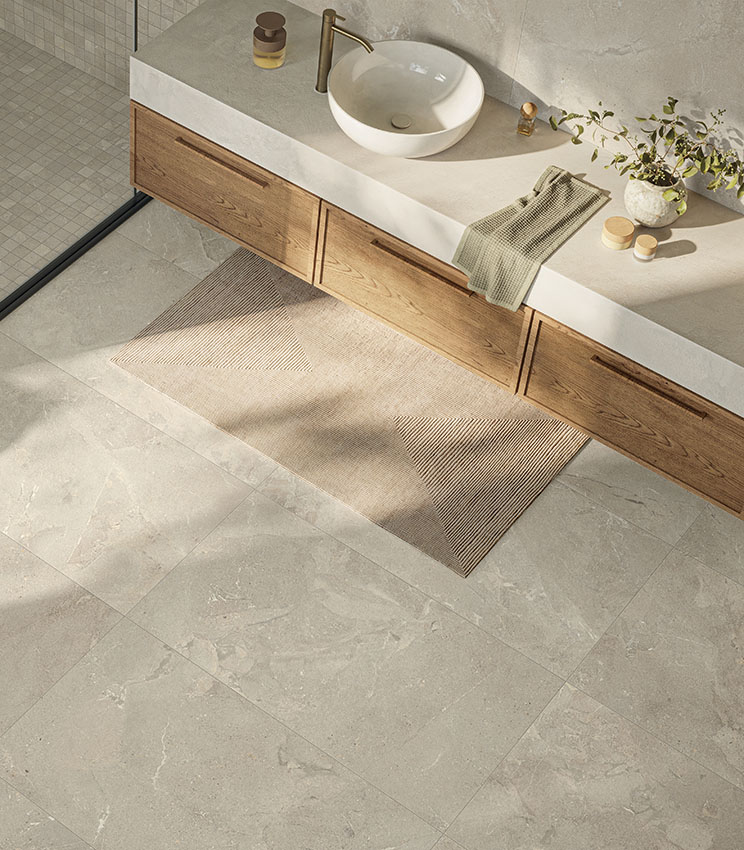 Salle de bains contemporaine avec des surfaces des collections Revel Dune, Mosaico Dune et Workshop Jade, caractérisée par un sol et des revêtements effet pierre dans des tons clairs. L’espace comprend un long meuble vasque en bois, un plan clair avec vasque blanche à poser, une robinetterie dorée, un tapis géométrique et une douche avec mosaïque coordonnée.